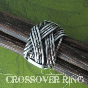 May include: A silver metal ring with a woven design. The ring is on a brown wooden surface.