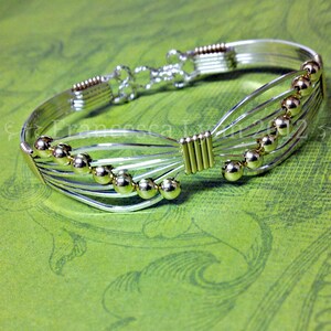 May include: A silver-toned wire bracelet with gold-colored accents and spherical beads. The bracelet features a unique design with multiple parallel wires and a decorative clasp. The gold accents are visible on the beads and at the center of the bracelet.
