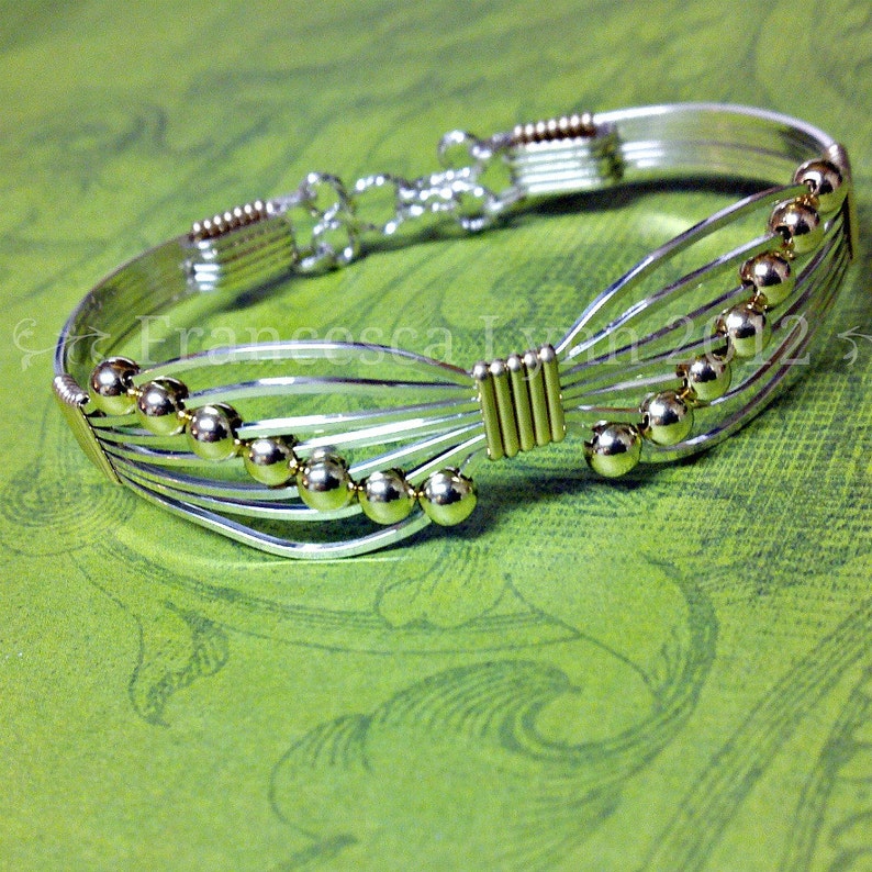 May include: A silver-toned wire bracelet with gold-colored accents and spherical beads. The bracelet features a unique design with multiple parallel wires and a decorative clasp. The gold accents are visible on the beads and at the center of the bracelet.