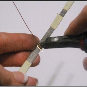 May include: Close-up of a person using pliers to cut a small piece of gold-colored wire. The wire is being worked on a silver-colored, tape-covered strip. A thin, brown wire extends from the gold piece.