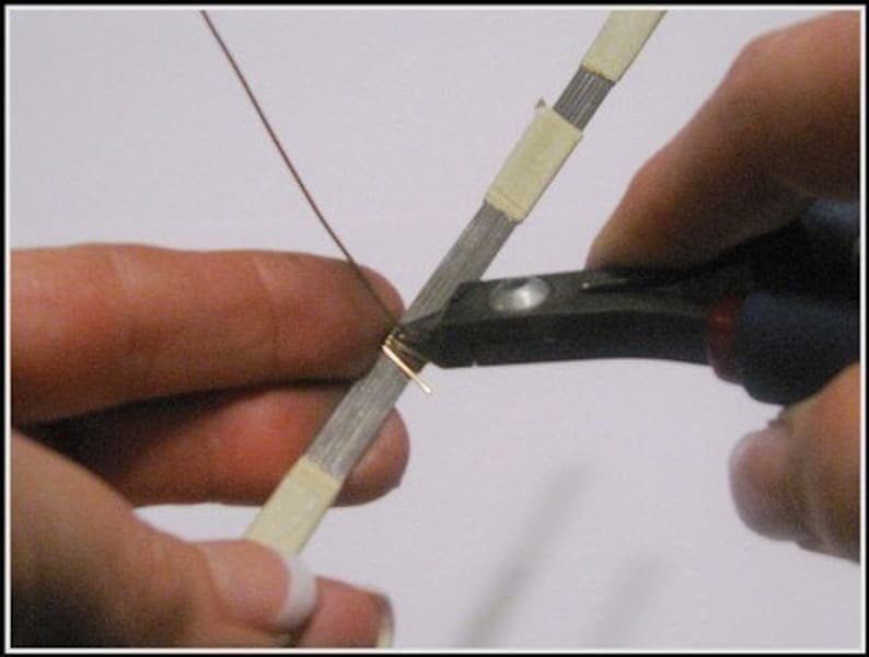 May include: Close-up of a person using pliers to cut a small piece of gold-colored wire. The wire is being worked on a silver-colored, tape-covered strip. A thin, brown wire extends from the gold piece.