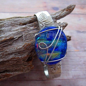 Puede incluir: Un colgante artesanal con una vibrante esfera de vidrio dicróico azul, envuelta intrincadamente en alambre plateado. El trabajo de alambre incluye espirales decorativas y una argolla para una cadena. El colgante se exhibe sobre un trozo de madera flotante.