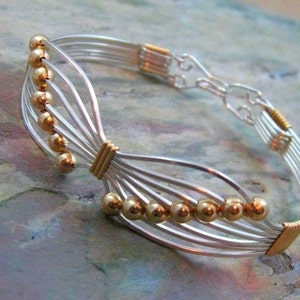 May include: A silver-toned wire bracelet with gold-colored accents. The bracelet features multiple strands of silver wire, interwoven with gold-colored beads and wrapped sections. The design is intricate and delicate, with a focus on the contrast between the silver and gold.