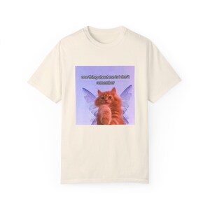 May include: Cream-colored t-shirt featuring a graphic of a ginger cat with purple fairy wings. The cat is holding its paw up. The graphic includes the text "one thing about me is I don't remember".