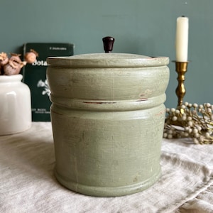 Può includere: Un contenitore vintage in legno verde chiaro con coperchio e pomello marrone scuro. Il contenitore cilindrico presenta scanalature orizzontali e vernice usurata. Si trova su una superficie di lino. Ideale per la decorazione.