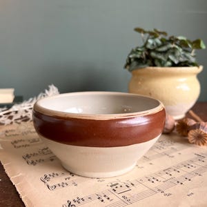 May include: A small, cream-coloured ceramic bowl with a wide, brown band around the middle. The bowl rests on a piece of vintage sheet music. A yellow pot with a green plant is in the background.