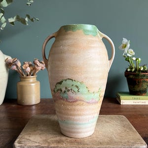 May include: A large, beige ceramic vase with two handles. The vase features green and pink painted accents. The vase is sitting on a wooden chopping board. The background is a dark teal wall.