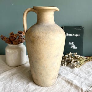 May include: A tall, beige ceramic pitcher with a handle. The pitcher has a slightly aged appearance, with subtle variations in color. A small white vase with dried seed pods and a book titled "Botanique" are in the background.