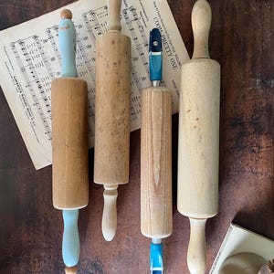 Può includere: Quattro mattarelli in legno di varie dimensioni e stili sono disposti su una superficie di legno scuro. Uno ha un manico azzurro, e un altro ha un manico blu e bianco. Sono visibili anche spartiti e un piccolo libro.