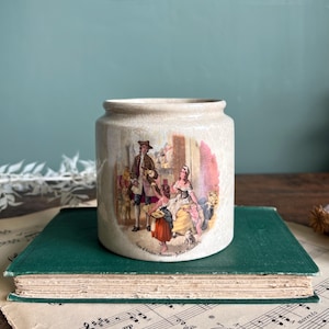 Può includere: Un vaso vintage in ceramica color crema con una dettagliata illustrazione di una scena storica. Il vaso è cilindrico con un bordo leggermente svasato. L'illustrazione raffigura figure in abiti d'epoca. Il vaso è appoggiato su un libro verde.