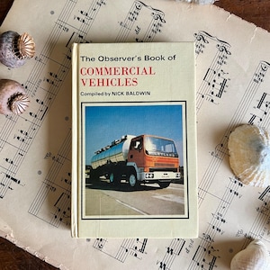 Könnte beinhalten: Ein Vintage-Buch mit dem Titel "The Observer's Book of Commercial Vehicles" von Nick Baldwin. Das Cover zeigt ein Farbfoto eines orange-weiß Tankwagens. Das Buch liegt auf einem Hintergrund aus alter Notenblätter und Muscheln.