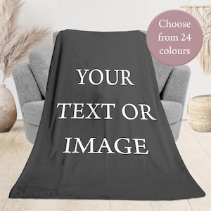 May include: A dark grey blanket draped over a grey armchair. The blanket has the white text "YOUR TEXT OR IMAGE". A circular graphic reads "Choose from 24 colours". Decorative items are in the background.