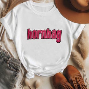 May include: A white short-sleeved t-shirt featuring the word "hornbag" printed in bold, pink, sans-serif letters with a black outline across the centre of the chest. The garment has a classic crew neckline and a standard fit with rolled sleeves.