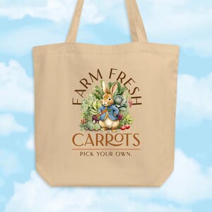 May include: Beige tote bag with a whimsical illustration of a rabbit in a blue jacket surrounded by vegetables. The text on the bag reads "FARM FRESH CARROTS PICK YOUR OWN."