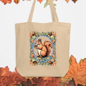 Può includere: Borsa tote beige con un'illustrazione incorniciata di uno scoiattolo che tiene una ghianda. Lo scoiattolo è circondato da un design floreale e di foglie nei toni dell'arancione, verde e blu. La borsa ha manici lunghi.