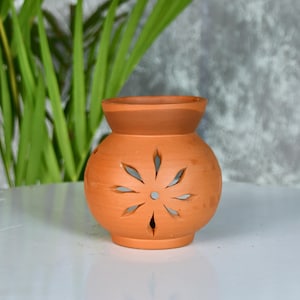 Puede incluir: Un difusor de aceites esenciales de terracota con un diseño recortado en forma de flor. La pequeña y redonda maceta de cerámica marrón anaranjado tiene una amplia abertura en la parte superior y se asienta sobre una superficie blanca. El fondo incluye plantas verdes y una pared gris.