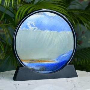 May include: A round sand art piece with a black frame and stand. The sand art features a blue and white sky with a golden-brown land and blue waves. The piece is displayed on a white marble surface.