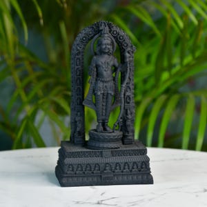 May include: A black statue of a deity, standing within an arched frame. The statue is intricately detailed, depicting a figure holding a bow and arrow. The base of the statue is rectangular with detailed carvings. The background features green foliage.