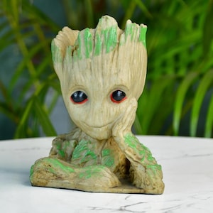 Puede incluir: Figura detallada de un personaje con apariencia de árbol, con tez marrón claro y acentos verdes. La figura tiene ojos grandes y expresivos con iris rojos y posa con una mano apoyada en la mejilla. La parte superior de la cabeza está abierta, lo que sugiere que es una maceta.