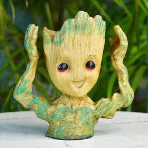 Puede incluir: Pequeña figura de maceta Groot sonriente. La figura es beige con detalles verdes en los brazos y la cabeza. Tiene grandes ojos negros con pupilas rojas. Los brazos están levantados y la cabeza está abierta para sostener una pequeña planta.