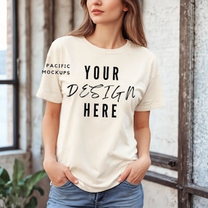 Puede incluir: Una mujer lleva una camiseta de color crema con el texto "YOUR DESIGN HERE" impreso en la parte delantera. La camiseta es un lienzo en blanco para un diseño personalizado.