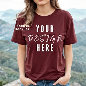 Op de afbeelding: Een maroon t-shirt met de tekst "YOUR DESIGN HERE" erop gedrukt. Het t-shirt wordt gedragen door een persoon die voor een bergketen staat.