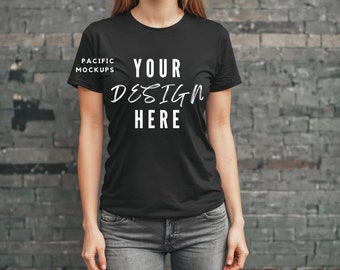 Puede incluir: Una camiseta negra con el texto "YOUR DESIGN HERE" impreso en la parte delantera. La camiseta la lleva puesta una persona de pie frente a una pared de ladrillo.