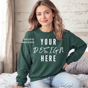 May include: A woman wearing a forest green sweatshirt with the text "YOUR DESIGN HERE" and "PACIFIC MOCKUPS" on the front. She is sitting on a bed with a white blanket.