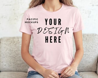 Puede incluir: Una camiseta rosa con el texto "YOUR DESIGN HERE" en la parte delantera. La camiseta la lleva puesta una persona sentada en un sofá.