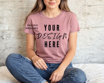 May include: A woman wearing a dusty rose t-shirt with the text "YOUR DESIGN HERE" printed on the front. She is sitting on a white surface with her legs crossed.