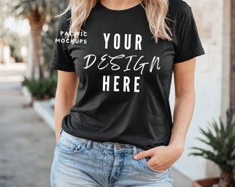 Puede incluir: Una camiseta negra con el texto "YOUR DESIGN HERE" en blanco. La camiseta la lleva puesta una persona con el pelo rubio y va combinada con unos pantalones cortos de mezclilla azul.