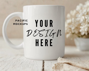 May include: White ceramic mug with the text "YOUR DESIGN HERE" and "PACIFIC MOCKUPS" on the side. The mug is sitting on a white wooden surface.