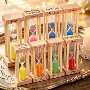 May include: A collection of eight wooden hourglasses with coloured sand, each featuring a name or phrase engraved on the top frame. The hourglasses contain yellow, pink, blue, green, orange, and red sand, arranged on a decorative metal tray.