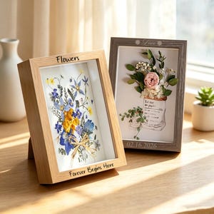 Peut inclure: Deux cadres vitrines rectangulaires. Le cadre de gauche en bois clair porte les inscriptions "Flowers" et "Forever Begins Here" avec des fleurs pressées bleues et jaunes. Le cadre gris porte "Love" et "02.16.2026" et contient une illustration florale.
