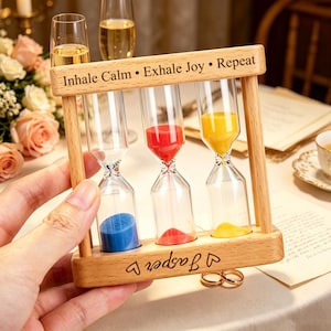 May include: A wooden frame holding three glass hourglasses with blue, red, and yellow sand. The top bar is engraved with the text Inhale Calm • Exhale Joy • Repeat, and the bottom bar features the engraved name Jasper with heart symbols on each side.