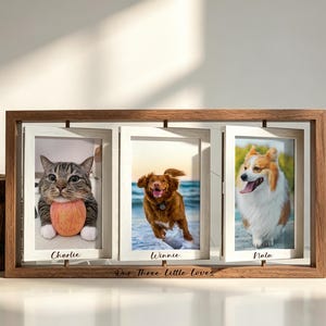 Op de afbeelding: Een rechthoekige houten lijst met drie draaibare fotolijsten. Elke lijst toont een foto van een huisdier met een naam eronder gedrukt: Charlie, Winnie en Nala. De basis van de houten lijst bevat de gegraveerde tekst Our Three Little Loves.