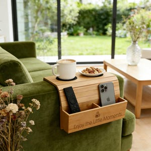 Puede incluir: Una bandeja de brazo de sofá de bambú con un portavasos, área para aperitivos y compartimentos para un mando a distancia y un teléfono. Contiene una taza de café blanca, un plato pequeño con nueces, un mando a distancia y un smartphone. El texto "Enjoy the Little Moments" está impreso.