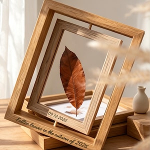 Puede incluir: Un marco flotante de madera marrón claro que contiene una hoja seca de color marrón. El marco presenta el texto grabado "Fallen leaves in the autumn of 2026" en el borde inferior y "09.12.2026" en el borde interior, con un panel de vidrio.