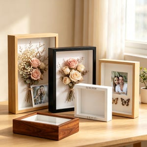 May include: A collection of rectangular shadow box frames in light wood, black, and white finishes. Two frames contain bouquets of dried roses and baby's breath with photos of a couple. One white frame features the text 02.16.2026 and S&K.