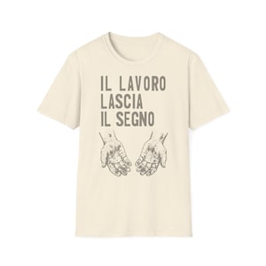 Può includere: Una t-shirt a maniche corte color crema con una grafica grigio scuro di due mani aperte e sporche di terra. Sopra le mani, il testo IL LAVORO LASCIA IL SEGNO è scritto in un carattere sans-serif in grassetto. Il tessuto ha una trama leggermente maculata.