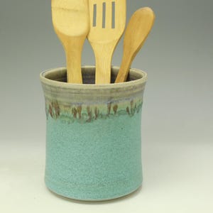 May include: A ceramic utensil holder in a speckled turquoise glaze with a light brown rim. Three wooden kitchen utensils are inside the holder. The holder is a cylindrical shape and sits on a white surface.