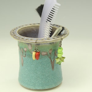 May include: A cylindrical ceramic holder with a turquoise and green drip glaze finish, featuring a wide rim with small indentations. A white plastic comb and two pairs of dangling earrings with colorful beads are stored inside the container.