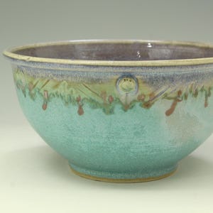 May include: A ceramic bowl with a rounded shape, featuring a turquoise and light blue glaze on the exterior. The upper rim displays a decorative band with subtle brown and green accents, while the interior has a smooth, deep purple finish.