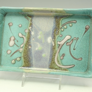May include: A rectangular ceramic tray with two side handles, featuring a turquoise glaze base with a central vertical band of light blue and green speckles, accented by abstract white and dark brown painted swirls and circular dots across the surface.
