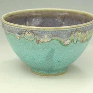 May include: A ceramic bowl featuring a vibrant turquoise lower section and a smooth, muted purple upper rim. The exterior surface displays a textured transition where the two colors meet, accented by small, raised, circular relief details along the edge.