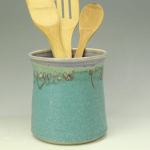 May include: A cylindrical ceramic utensil holder with a speckled turquoise glaze and a lavender rim. The exterior features a decorative drip pattern in shades of brown and green. Three light-colored wooden kitchen utensils are placed inside.
