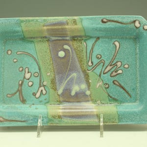 May include: A rectangular ceramic tray with a turquoise base and a decorative design. The tray features a central purple stripe, flanked by green stripes, and adorned with white and brown abstract accents. The tray is supported by a clear stand.