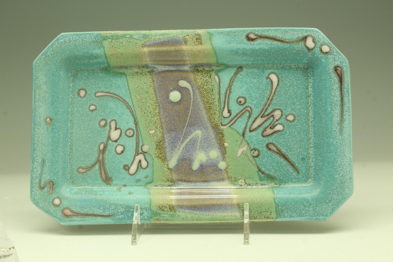 Puede incluir: Una bandeja rectangular de cer&aacute;mica con base turquesa y dise&ntilde;o decorativo. La bandeja presenta una franja central morada, flanqueada por franjas verdes, y adornada con detalles abstractos blancos y marrones. La bandeja est&aacute; sostenida por un soporte transparente.