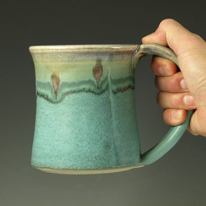May include: A ceramic mug with a flared rim and a sturdy handle. The body features a textured, speckled finish in shades of teal and turquoise, transitioning to a lighter, creamy green glaze near the top with subtle, dark brown drip accents.