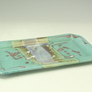 May include: A rectangular ceramic tray featuring a light turquoise glaze with a central band of blended blue, purple, and brown tones. The surface is decorated with scattered abstract splatters in dark pink and white, creating a textured appearance.